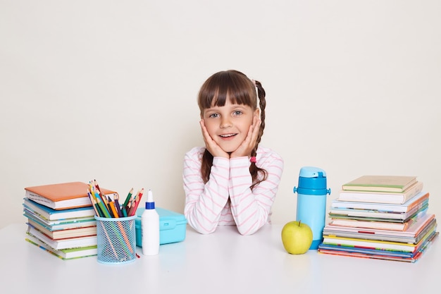 Portrait of smiling pretty cute little schoolgirl with dark hair and braids sitting at table surrounded with books looking at camera and keeps palms on cheeks being in good mood