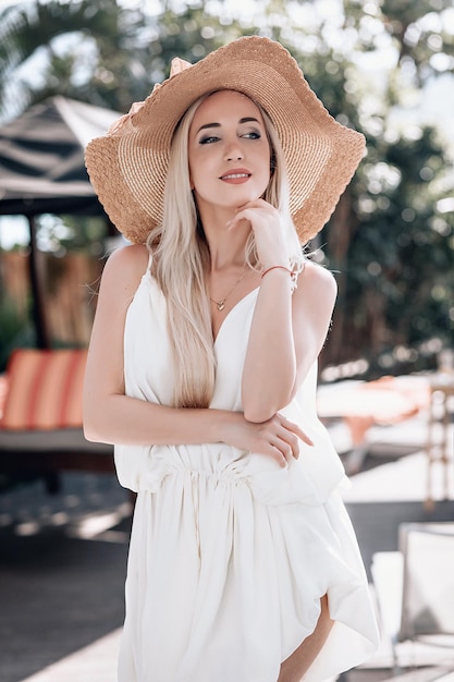Portrait of smiling pretty female with evening make up in a fashion white dress and a big  straw hat standing and a posing at the camera in a hotel resort. Phuket. Thailand