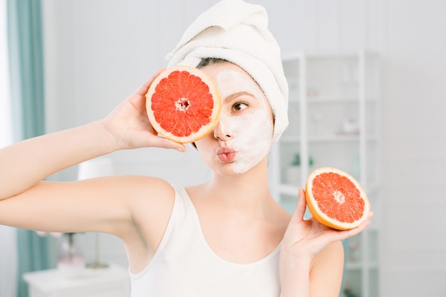 Portrait of smiling pretty girl with white towel on head holding halves of grapefruits near face closing one eye, healthy perfect smooth skin, white mask on one half of face, over light space