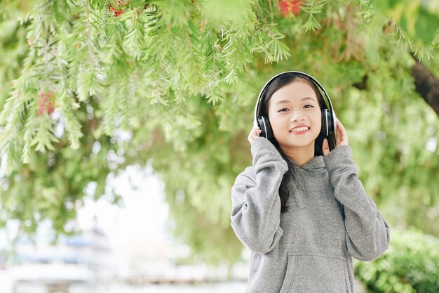 Portrait of smiling pretty Vietnamese girl listening to music in headphones when spending day in park