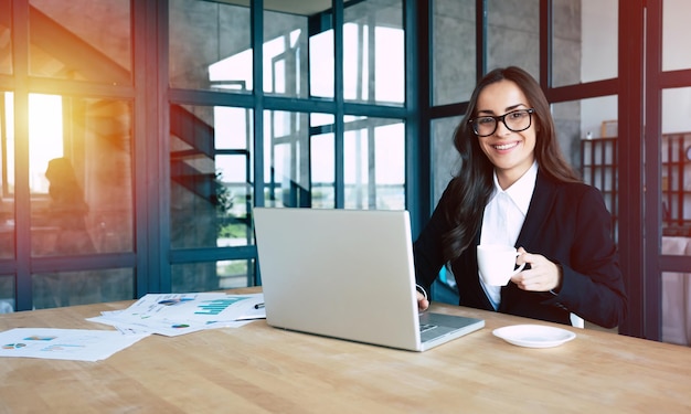 Portrait of smiling pretty young business woman in glasses and full suit sitting on workplace at office