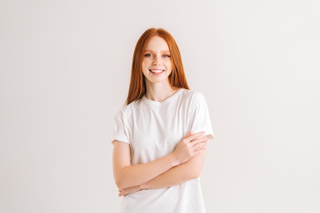 Портрет улыбающейся красивой молодой женщины, смотрящей в камеру, стоя со скрещенными руками на белом