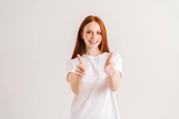 Portrait of smiling pretty young woman showing double thumbs up looking at camera standing on white