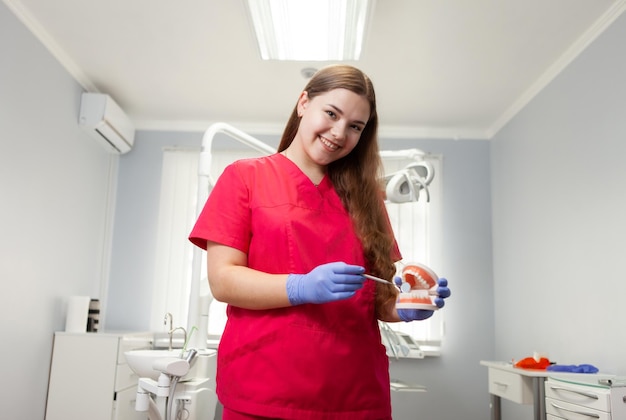 Portrait of smiling professional female dentist in red medical uniform at dentist office