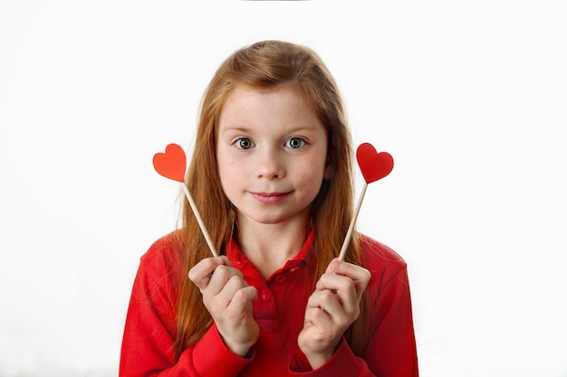 Foto ritratto della bambina caucasica dai capelli rossi sorridente che tiene i cuori rossi sui bastoni nelle sue mani. amore, san valentino, festa della mamma, festa del papà o concetto di buon compleanno.