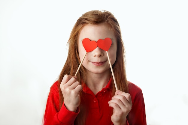 Портрет улыбающейся рыжеволосой маленькой девочки, закрывающей глаза красными сердечками на палочках.