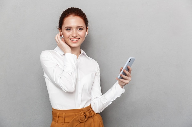 Портрет улыбающейся рыжей деловой женщины 20-х годов в наушниках, держащей и смотрящей на смартфон в офисе, изолированной над серым