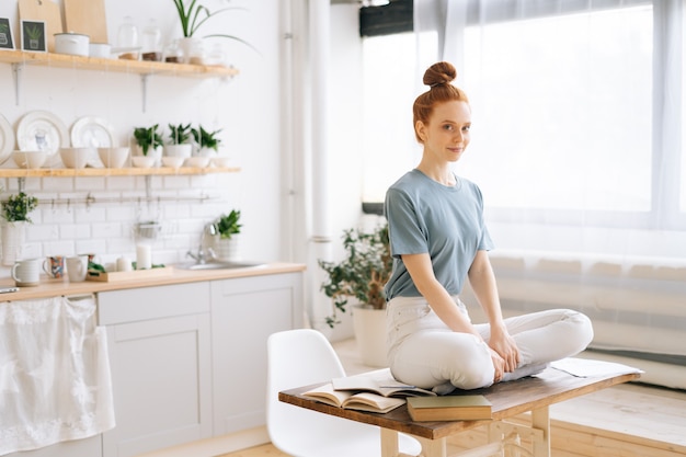 Portrait of smiling redhead young woman meditating while sits on desk at home office