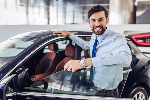 Portrait of smiling salesman standing at the car with opened door in dealership salon and looking at the camera