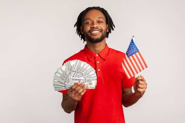 Portrait of smiling satisfied man holding american flag and dollars banknotes, celebrating success.