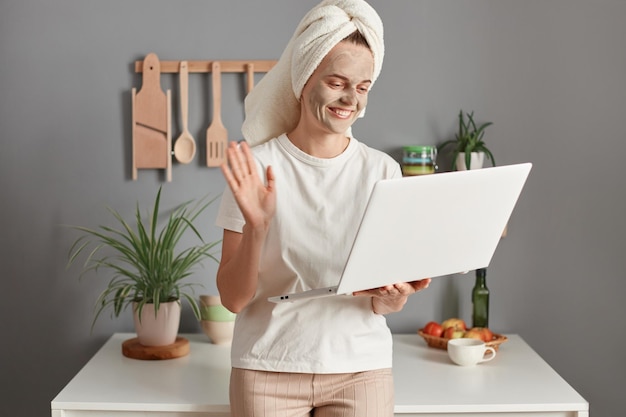 Portrait of smiling satisfied woman wearing white Tshirt and wrapped in towel with beauty treatment facial mask using her laptop for having video call waving hand saying hello