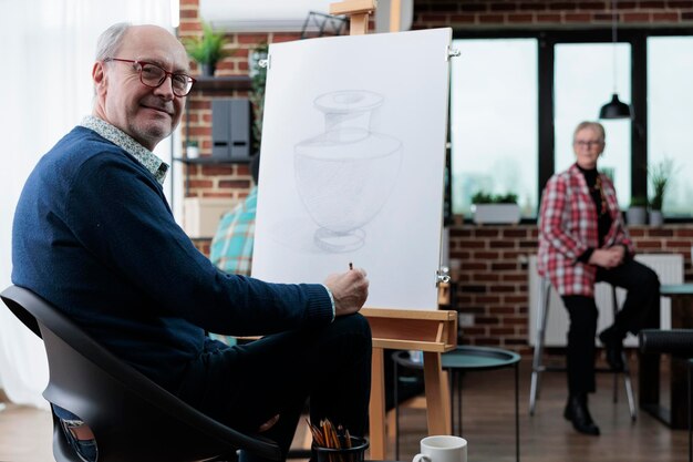 Ritratto di artista senior sorridente in piedi di fronte su tela bianca che disegna un vaso artistico utilizzando la tecnica di schizzo durante la lezione d'arte nello studio della creatività. pittore anziano che ha un hobby creativo