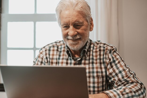 Portrait of smiling senior bearded man sitting at table with laptop browsing online sites Elderly man in checkered shirt using new technologies by shopping or watching videos