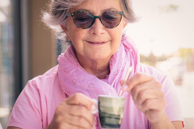 Portrait of smiling senior caucasian woman in pink and sunglasses holding a coffee cup sitting outdoor in a coffee shop