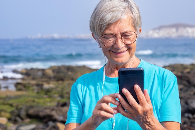 이어폰으로 듣고 있는 매력적인 노부인 전화를 사용하여 해변에 서 있는 웃고 있는 백인 노부인의 초상화