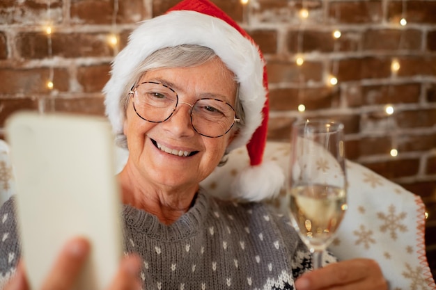 Portrait of smiling senior caucasian woman in winter sweater\
sitting at home holding a champagne flute while using phone video\
chatting with family or friends celebrate christmas and new\
year