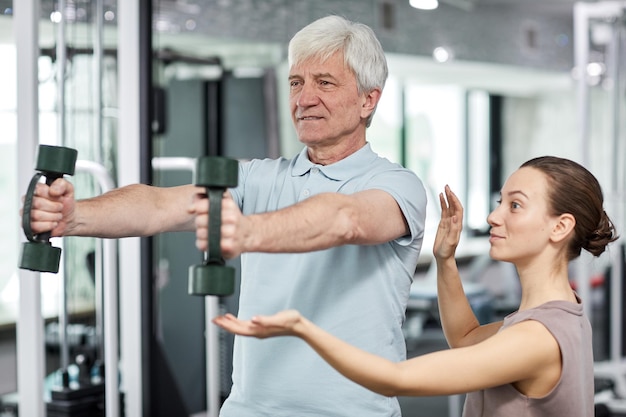 Portrait of smiling senior man doing exercises in gym at rehabilitation clinic with female therapist