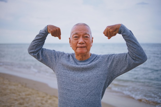 Photo portrait of smiling senior man flexing muscles while standing at beach against sky during sunset