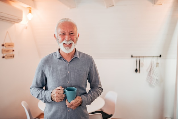 Photo portrait of a smiling senior man standing in the bright room, holding a mug.