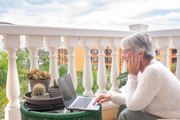 Портрет улыбающейся пожилой женщины, просматривающей ноутбук с домашнего балкона, взрослой привлекательной пожилой женщины, наслаждающейся тихим образом жизни на открытом воздухе, работающей за компьютером