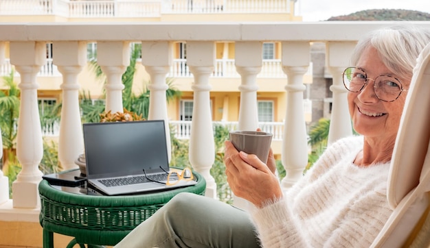 Портрет улыбающейся пожилой женщины, отдыхающей дома на балконе с чашкой кофе, смотрящей в камеру Взрослые привлекательные женщины, наслаждающиеся тихим образом жизни на открытом воздухе