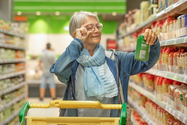 Портрет улыбающейся пожилой женщины, делающей покупки в супермаркете, выбирающей проверку продукта Кавказский пожилой покупатель в продуктовом магазине с тележкой для покупок