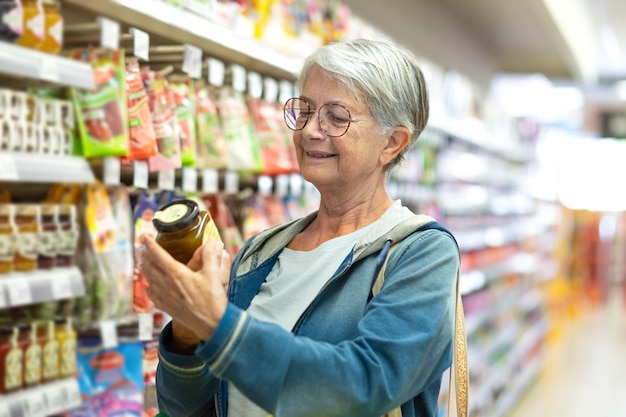 Portrait smiling senior woman making purchases in the supermarket selecting some jam Caucasian elderly customer in grocery store