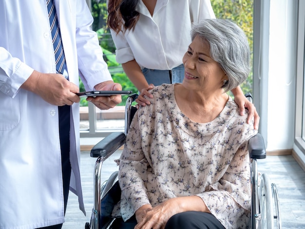 Portrait of smiling senior woman patients in wheelchair with assistant take her to meet doctor male use tablet discussing her medical treatment records Senior adult checkup appointment concept