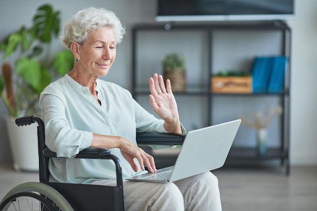 Portrait of smiling senior woman in wheelchair waving at camera while using video chat at home copy ...