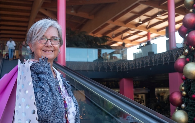 Ritratto di una donna anziana sorridente dai capelli bianchi che fa shopping per natale in un centro commerciale, mentre tiene in mano molte borse con regali per la famiglia e gli amici. il concetto di consumismo