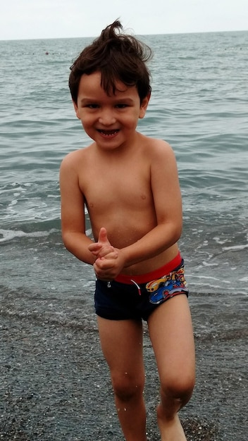 Portrait of smiling shirtless boy walking at beach against sky