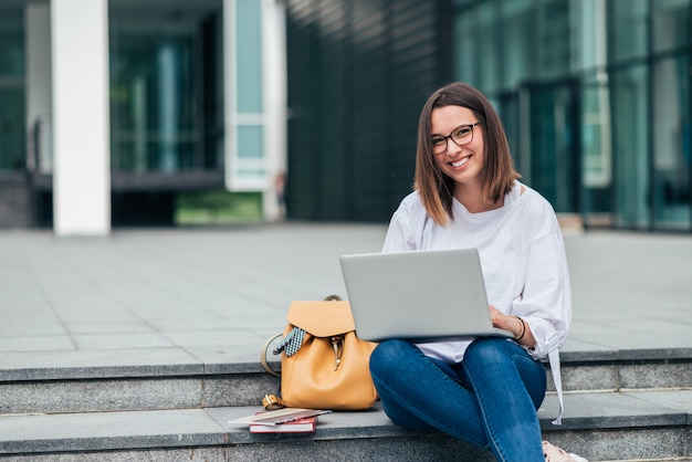 Portrait of a smiling student girl sitting on stairs in the city and using laptop.