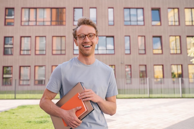 Photo portrait of smiling student holding book and laptop looking at camera standing in university campus