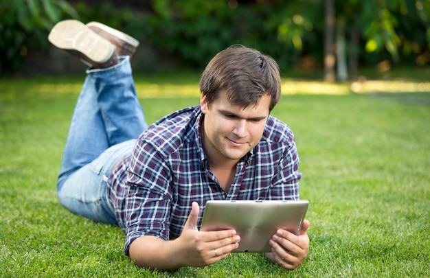 Портрет улыбающегося студента с помощью цифрового планшета на траве в парке