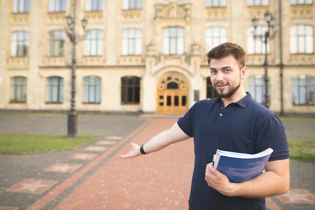 Foto ritratto di uno studente sorridente con libri e quaderni nelle sue mani in piedi all'ingresso dell'università e invita con la mano