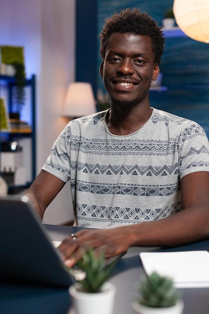 Portrait of smiling student working remote from home at business project using laptop computer. Young freelancer sitting at desk in living room during online financial lesson. Distance education