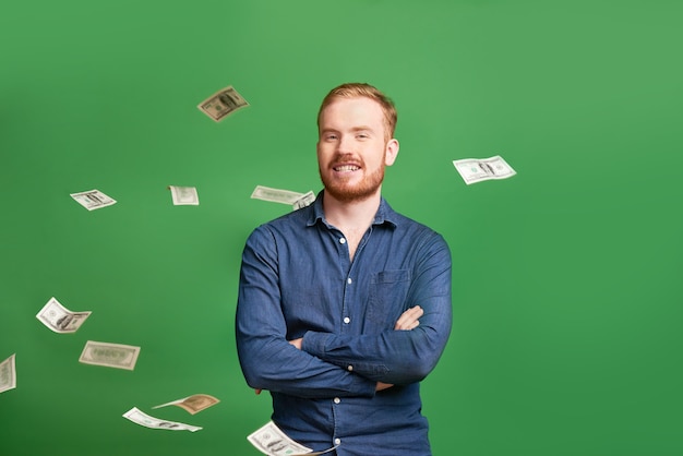 Portrait of smiling successful young redhead male entrepreneur with crossed arms standing under falling banknotes