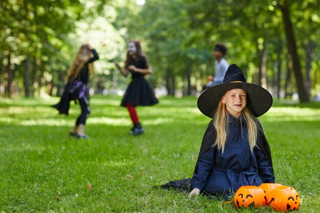 Ritratto dell'adolescente sorridente vestito come strega per halloween che si siede sull'erba verde all'aperto con i bambini che giocano in superficie, lo spazio della copia