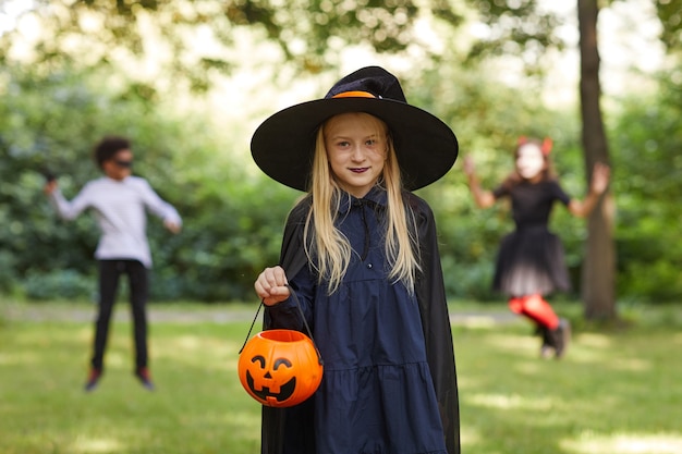 Portrait of smiling teenage girl dressed as witch posing outdoors and holding Halloween bucket with kids playing in surface, copy space