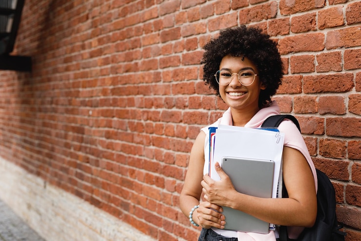 Photo portrait of smiling teenage girl holding books against brick wall
