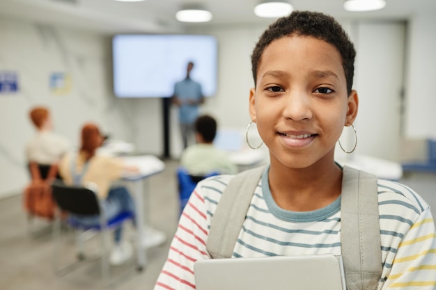 Photo portrait of smiling teenage girl holding books and looking at camera in school classroom, copy space