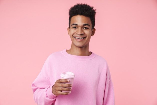 Portrait of a smiling teenager boy wearing pullower standing isolated over pink wall, holding takeaway coffee cup