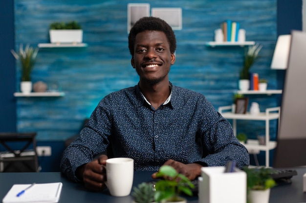 Portrait of smiling teenager holding cup of coffee while studying at management lesson using online university class during coronavirus lockdown. Black man working remote from home