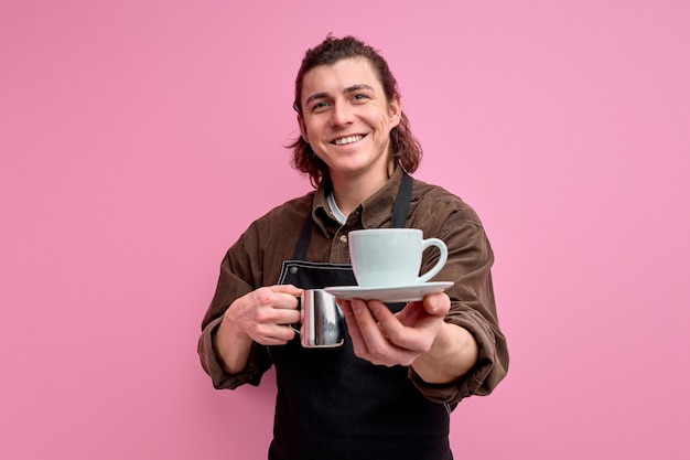 Portrait of smiling waiter guy student work part-time in coffee bar shop give to client mug of coffee