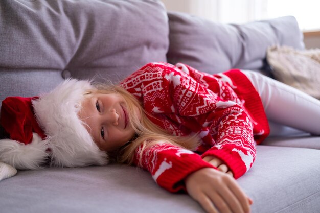 Portrait smiling waiting for holidays girl with in santa hat and christmas costume sitting at home