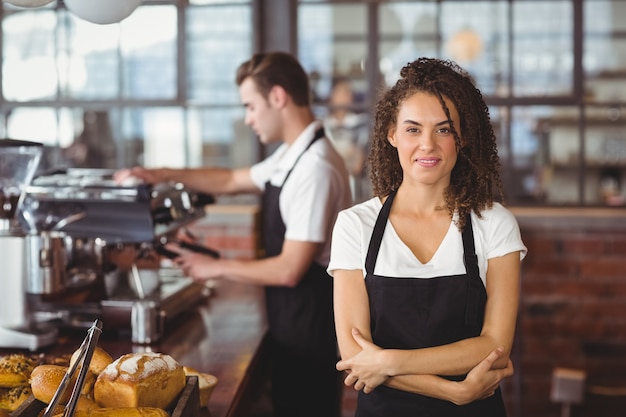 Portrait of smiling waitress with arms crossed in front of colleague at coffee shop