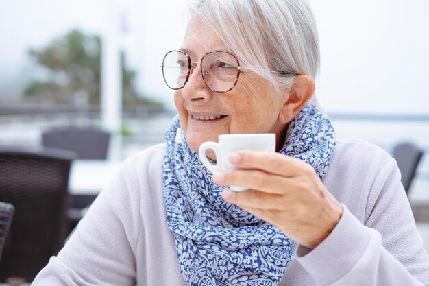 Portrait of smiling white haired senior woman sitting outdoors at cafe table holding a coffee cup