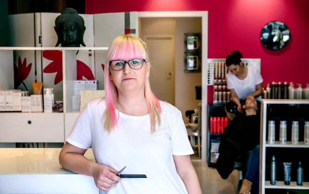 Portrait of smiling woman hairdresser standing in a hair and beauty salon with employee washing woman hair in the background