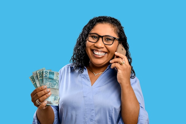 Portrait of smiling woman holding 100 reais banknotes talking on cellphone isolated on blue background