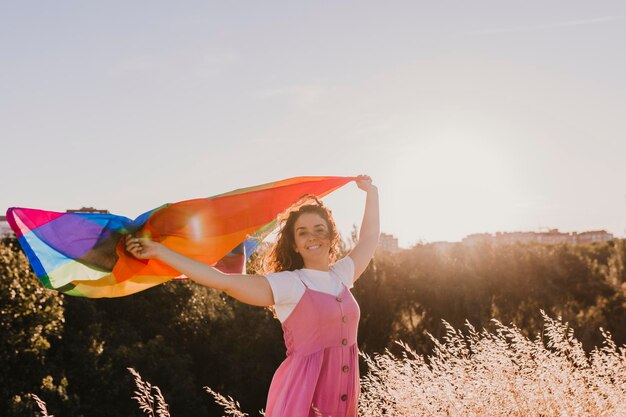 Photo portrait of smiling woman holding rainbow flag while standing on field during sunset